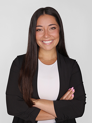 A woman with long dark hair, wearing a black blazer over a light top, smiles confidently with her arms crossed against a plain light background.