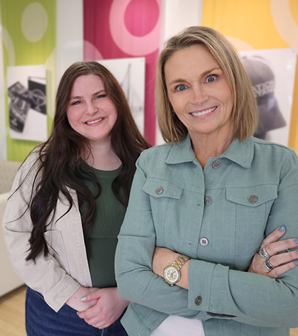 Two women smile at the camera in a brightly lit room with colorful, patterned banners and black-and-white photos on the wall behind them. One stands in front with arms crossed; the other stands slightly back.