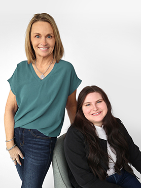 Two women pose together against a white background. One stands smiling with hands in her pockets, wearing a teal top and jeans. The other sits and smiles, with long dark hair, wearing a black jacket and white shirt.