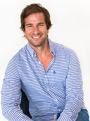 A man with brown hair is smiling and sitting against a white background. He is wearing a blue and white checkered shirt with the top buttons undone and blue jeans.