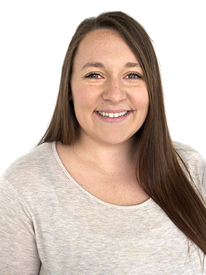 A woman with long brown hair and a light beige top smiles at the camera against a plain white background.