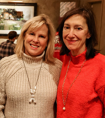 Two women smiling and standing close together indoors. One wears a cream-colored sweater with a long necklace, and the other wears a bright red sweater with a gold chain. A painting and other people are in the background.
