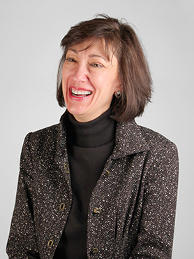 A woman with short brown hair, wearing a black turtleneck and a textured dark blazer, smiles and looks slightly to her left against a plain light background.