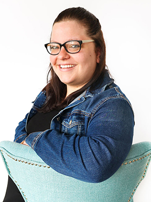 A smiling woman with long dark hair and glasses sits sideways in a light blue studded chair, wearing a denim jacket over a black shirt, against a plain white background.