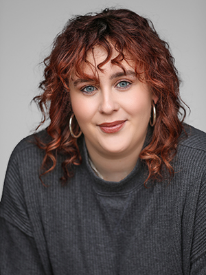 Woman with wavy, shoulder-length auburn hair and blue eyes, wearing large hoop earrings and a dark gray ribbed sweater, smiles softly at the camera against a neutral background.