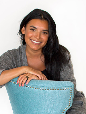 A woman with long dark hair and a warm smile sits behind a light blue chair with silver studs. She wears a gray cardigan and rests one arm on the chair, posing against a plain white background.