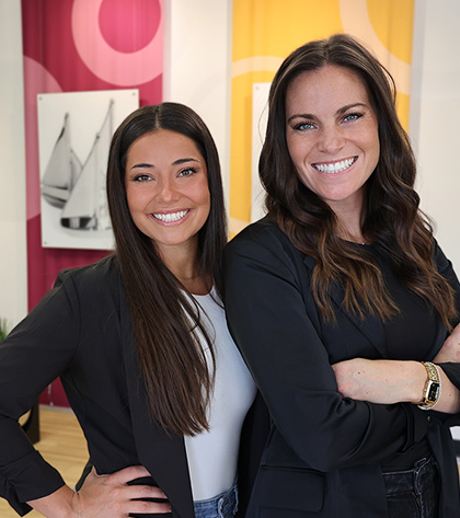 Two women standing side by side indoors, smiling at the camera. Both have long hair and are wearing black jackets. Colorful, modern artwork and bright yellow and pink panels are visible in the background.