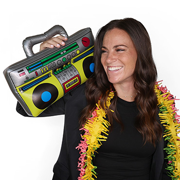 Smiling woman with long brown hair wears a colorful lei and holds an inflatable boombox prop on her shoulder, posing against a plain white background.