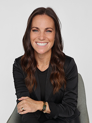 A woman with long brown wavy hair, wearing a black blazer and top, sits on a gray chair and smiles at the camera against a plain light background.