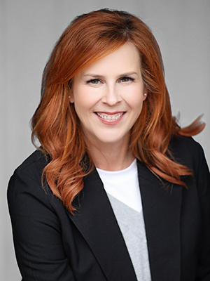 Smiling woman with long, wavy red hair wearing a black blazer over a white and gray top, posed against a plain gray background.