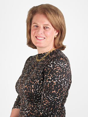 A woman with shoulder-length light brown hair, wearing a black, brown, and white patterned top and a gold necklace, smiling at the camera in front of a plain white background.