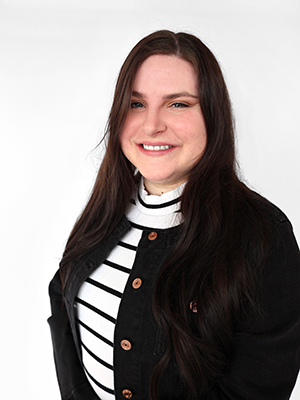 A woman with long dark hair smiles at the camera. She is wearing a black jacket over a white turtleneck with black horizontal stripes, standing against a plain white background.