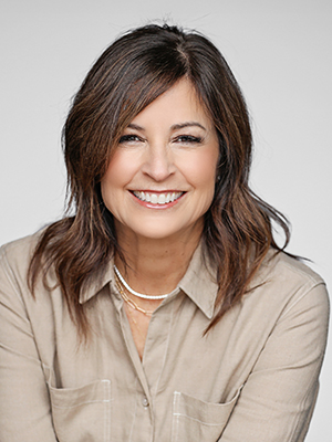 A woman with shoulder-length brown hair, wearing a beige button-up shirt and layered necklaces, smiles warmly at the camera against a light gray background.