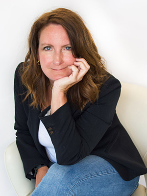 A woman with wavy brown hair, wearing a black blazer, white shirt, and blue jeans, sits on a white chair. She rests her chin on her hand and looks directly at the camera with a slight smile.