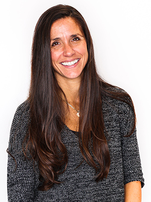 A woman with long brown hair wearing a gray sweater smiles at the camera against a plain white background.