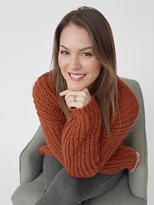 A woman with straight brown hair, wearing a chunky orange sweater and dark jeans, sits in a light gray chair, smiling at the camera with her chin resting on her hand.
