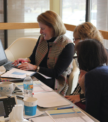 Three women sit at a table covered with papers, a laptop, coffee cups, and a water bottle. They are smiling and working together, appearing engaged and collaborative in an office setting with large windows.