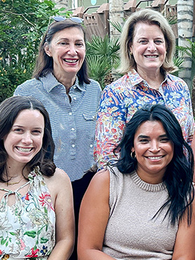 Four women are smiling together outdoors. Two are standing in the back, wearing patterned shirts, and two are seated in front, wearing sleeveless tops. Green plants and trees are visible in the background.