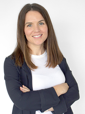 A woman with straight brown hair wearing a white top and a dark blazer stands smiling with her arms crossed against a plain white background.