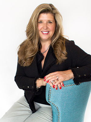 A woman with long, wavy blond hair sits on a blue studded chair. She is wearing a black jacket, gray pants, and has red-painted nails. She is smiling and looking at the camera against a white background.
