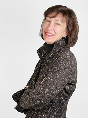 A woman with short brown hair wearing a textured black coat, smiling and standing with arms crossed against a plain white background.