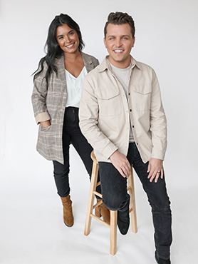 Two people posing in a studio: a woman with long dark hair in a plaid blazer standing, and a man with short hair in a light shirt and black jeans sitting on a wooden stool. Both are smiling at the camera.