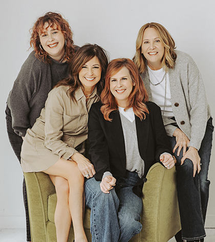 Four women pose together, smiling. Two sit on a green armchair, while the other two stand behind them. They all have shoulder-length hair in shades of red or light brown and wear casual outfits in neutral colors.