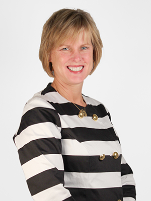 A woman with short blonde hair smiles at the camera. She is wearing a black and white striped jacket with large buttons and a necklace, standing against a plain white background.