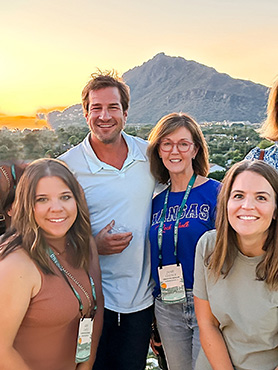 A group of four smiling adults, two men and two women, pose outdoors at sunset with a mountain in the background. They are wearing casual clothes and conference badges.