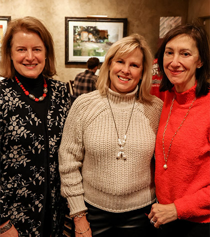 Three women stand together indoors, smiling at the camera. They are dressed in winter clothing, including sweaters and necklaces. Framed artwork hangs on the wall behind them.