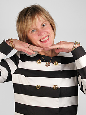 A woman with short blonde hair smiles playfully, resting her chin on her hands. She wears a black and white striped jacket with gold buttons and jewelry, standing against a plain light background.