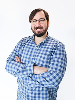 A man with glasses and a beard, wearing a blue plaid shirt, stands smiling with his arms crossed against a plain white background.