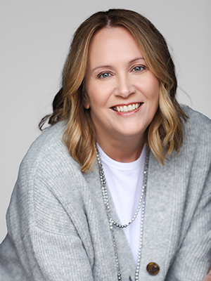 A woman with shoulder-length blonde hair smiles at the camera. She is wearing a light gray cardigan over a white top and a long beaded necklace, with a neutral background.