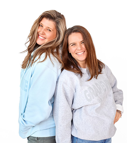Two women stand back to back, both smiling at the camera. They are wearing casual sweatshirts; one is light blue and the other is light gray. The background is plain white.