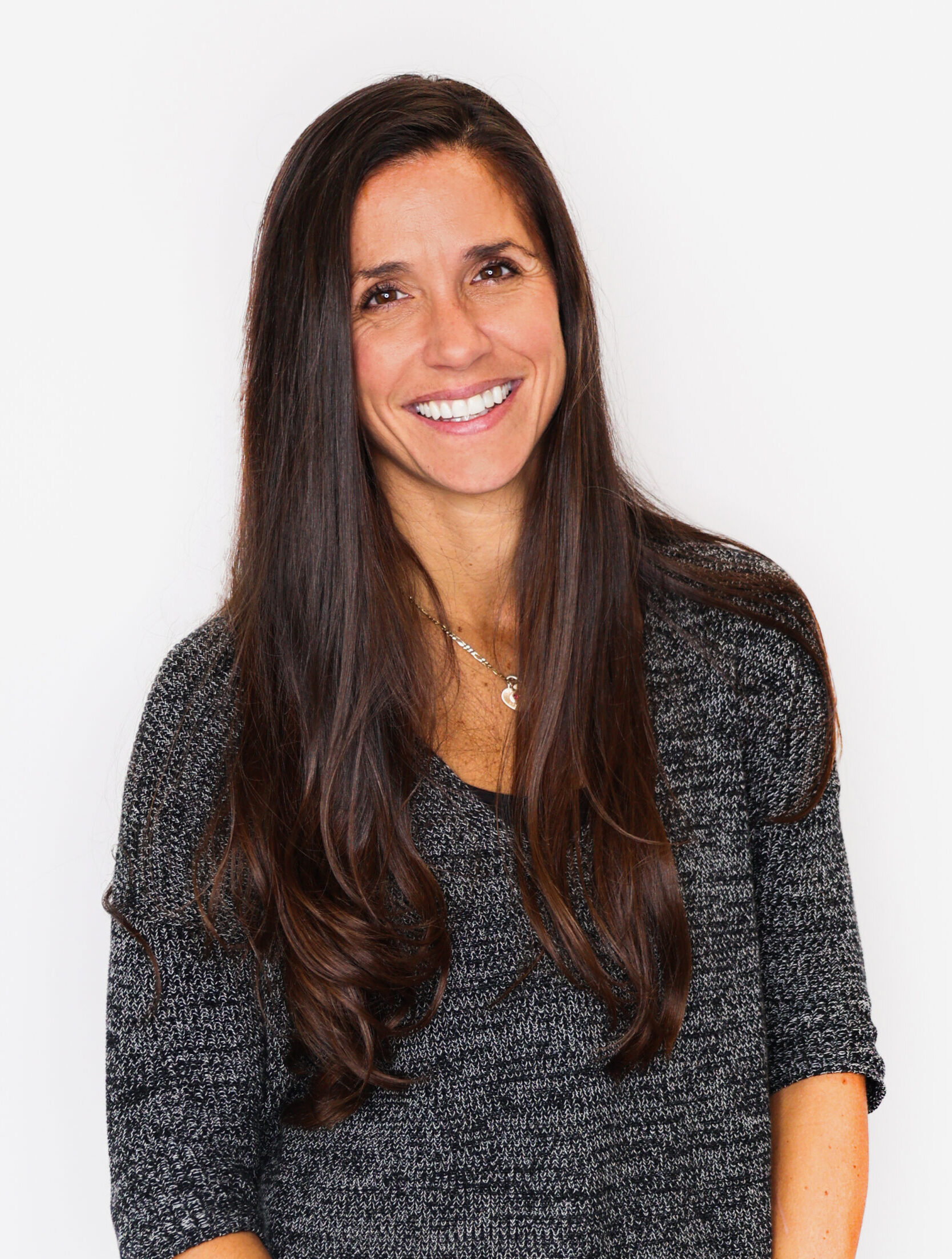 A woman with long brown hair wearing a gray knit sweater stands against a white background, smiling warmly at the camera with her arms relaxed by her sides.