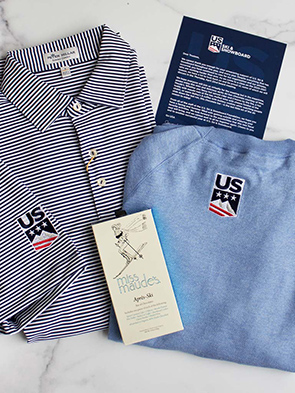 Two shirts with US Ski & Snowboard logos, one blue striped and one solid light blue, are displayed on a marble surface alongside a skincare box and an informational card—perfect for a promotional marketing agency’s next campaign.