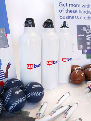 Three white US Bank water bottles, US Bank-branded stress balls shaped like baseballs and footballs, and pens are arranged on a table, showcasing the work of a top Promotional Marketing Agency with vibrant promotional signs in the background.