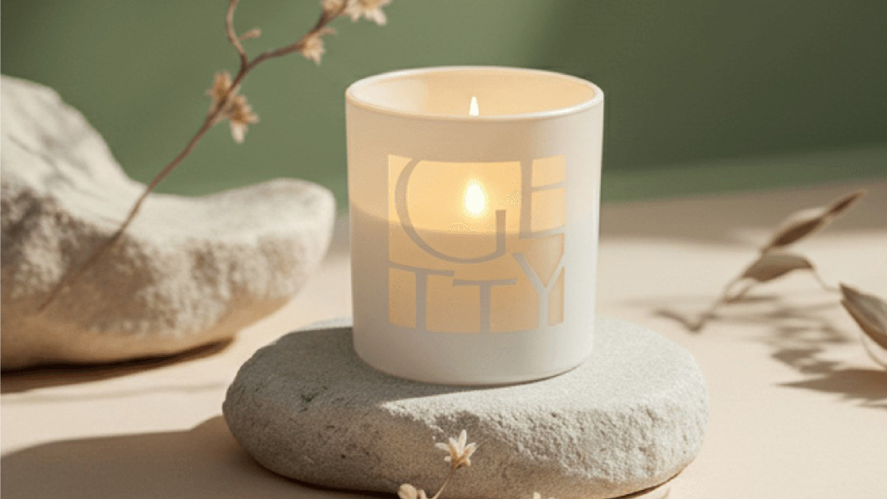 A white candle in a glass holder with the word “GETTY” sits lit on a smooth stone, surrounded by dried plants and a green background—a calming piece of health and wellness branded merchandise designed to inspire tranquility in any setting.
