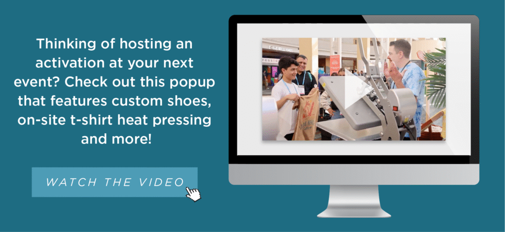 A computer screen displays an image of a pop-up event with two people interacting at a t-shirt heat pressing station. Text beside the monitor promotes custom shoes, on-site t-shirt pressing, and a video to watch.