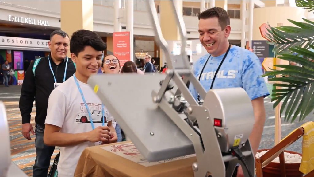 Several people smile and laugh together while using a large heat press machine at an indoor event. The setting appears lively, with visible signage and hall booths in the background, as participants enjoy crafting bespoke experiences together.