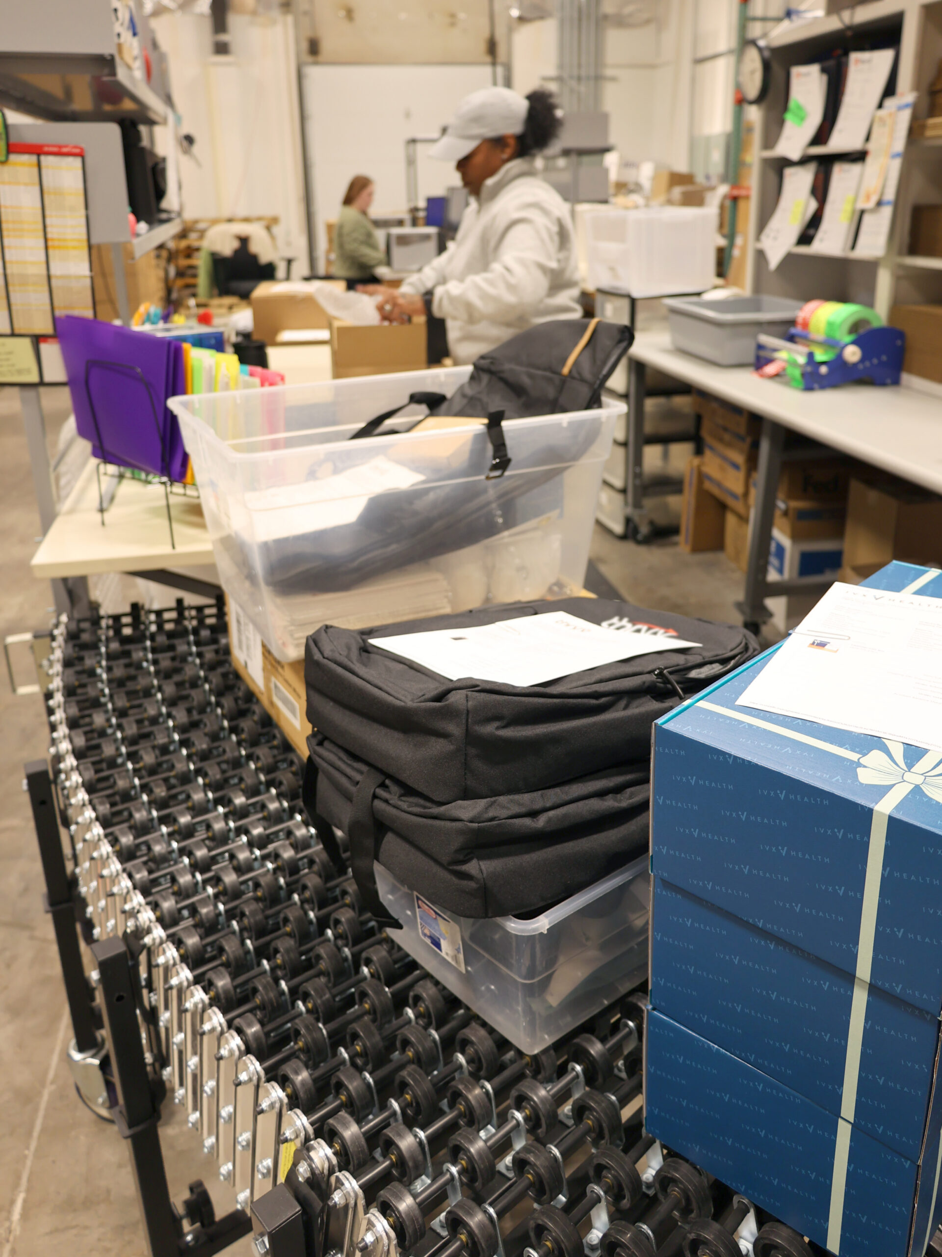 A person works at a packing and kitting station surrounded by shelves, paperwork, and plastic bins. Boxes, folders, and black bags are placed on a roller conveyor in the foreground.