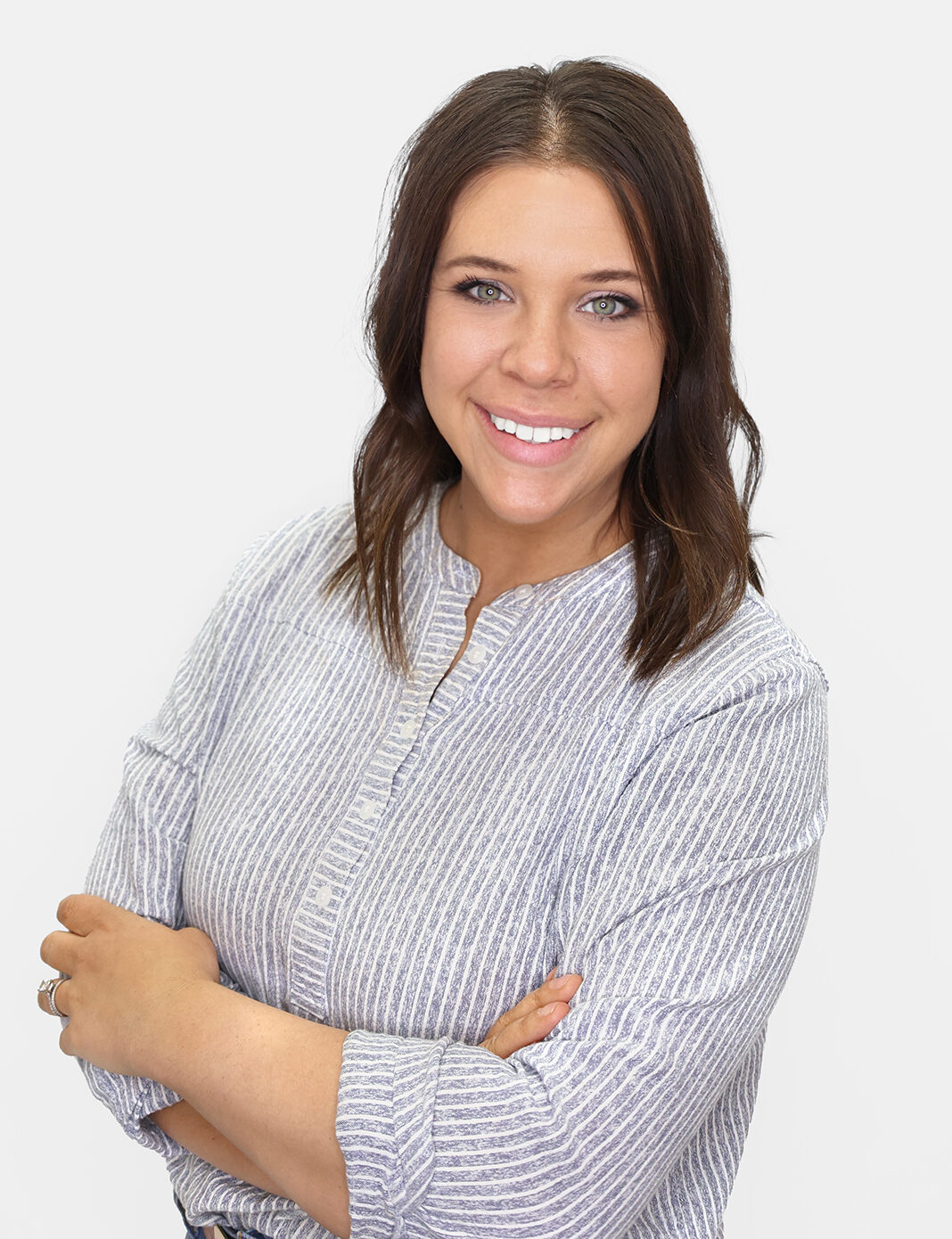 A woman with shoulder-length brown hair and blue eyes smiles at the camera. She is wearing a light grey and white striped blouse and has her arms crossed against a plain white background.
