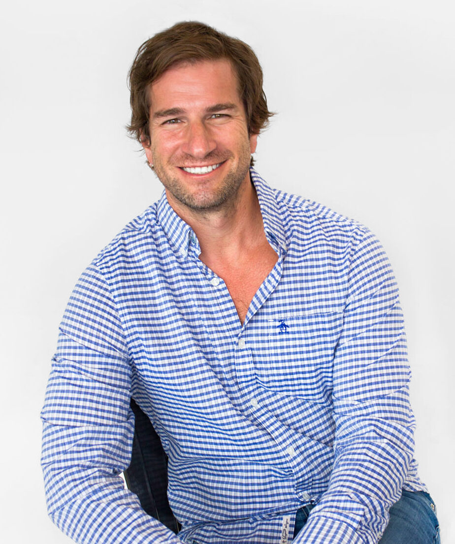 A man with brown hair wearing a blue and white checkered shirt sits and smiles against a plain white background.