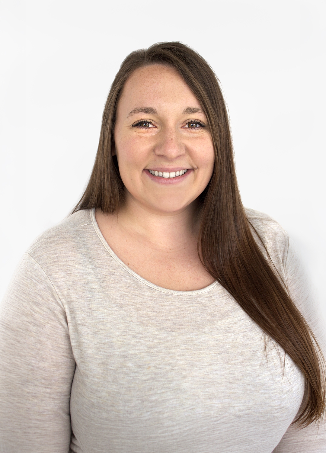 A woman with long brown hair and a light beige top smiles at the camera against a plain white background.