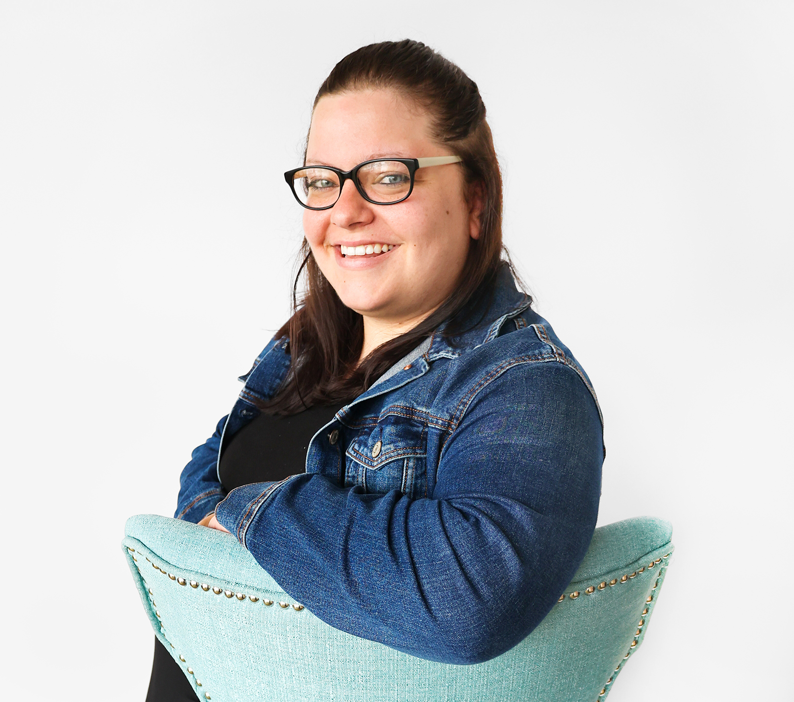 A woman with long brown hair, wearing glasses, a black shirt, and a blue denim jacket, smiles while sitting sideways on a light blue chair with silver studs against a plain white background.