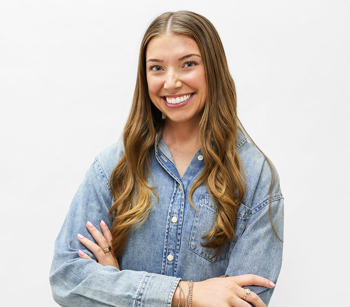 A woman with long brown hair, wearing a denim button-up shirt and black pants, stands smiling with her arms crossed against a plain white background.