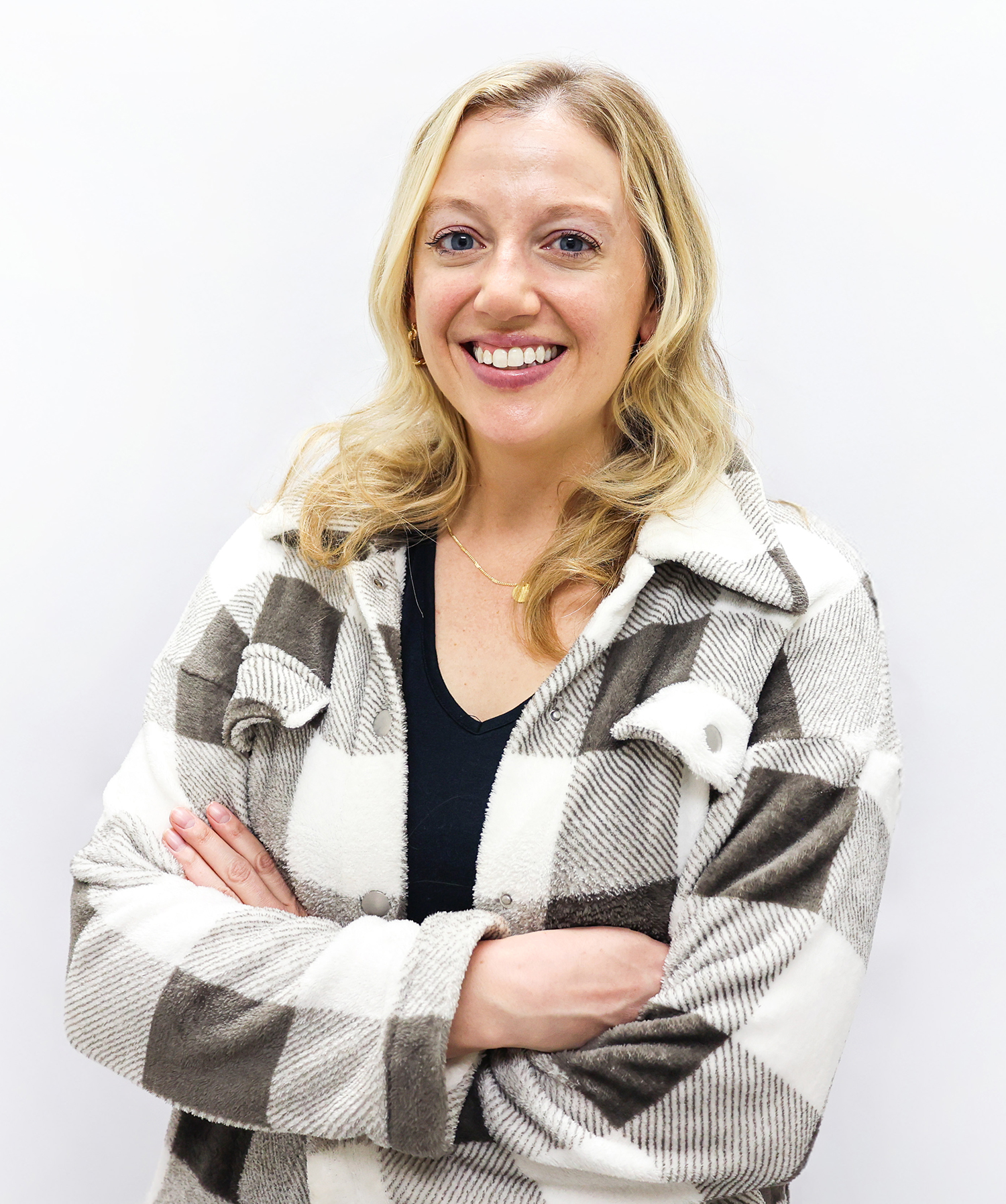 A woman with blonde hair and a big smile stands against a plain white background, wearing a black top and a cozy, checkered jacket with her arms crossed.