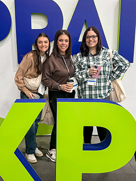 Three smiling women stand together indoors, holding drinks and wearing name badges. They pose in front of large, colorful letters on a wall, with a bright green "K" in the foreground.