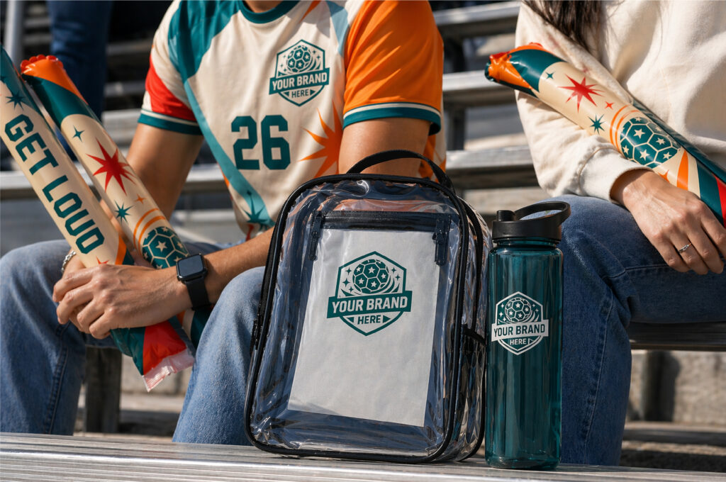 Two people in branded sports jerseys sit on bleachers holding colorful "Get Loud" noisemakers. A clear backpack and a water bottle, both featuring the same logo, are placed in front of them.