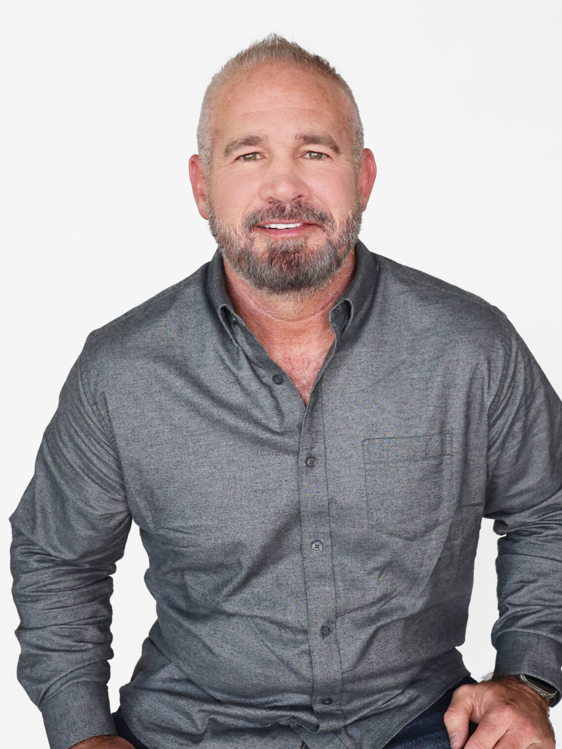 A man with short gray hair and a beard sits on a wooden stool, wearing a gray button-up shirt and dark jeans, smiling confidently at the camera—perfect for a promotional marketing company’s campaign against a plain white background.